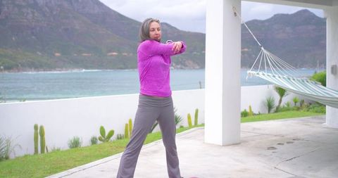 Mature woman stretching on coastal patio overlooking ocean wearing purple top