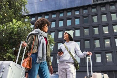 Two women walking and chatting with rolling luggage on urban sidewalk near glass building