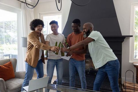 Diverse male friends clinking beer bottles in cozy living room toasting celebration