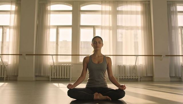 Woman meditating on yoga mat in sunlit studio with arched windows and ballet barre