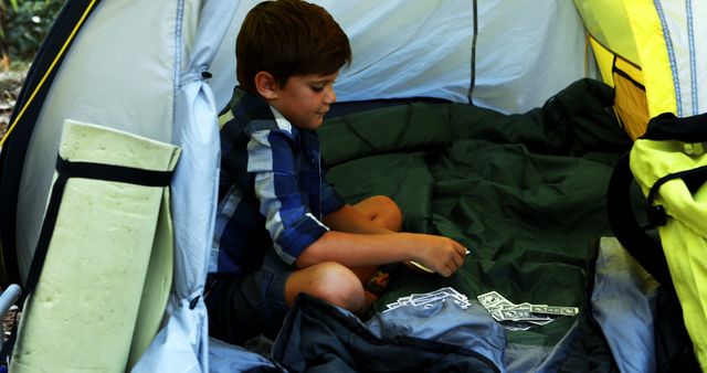 Young boy in a camping tent playing solitaire with cards. Ideal for themes of outdoor activities, children's leisure time, nature, and camping adventures. Use in content related to camping tips, family vacations, and promoting outdoor activities for children.