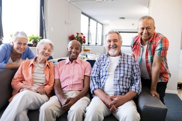 Group of diverse senior individuals sitting on a couch in a nursing home, smiling and enjoying each other's company. Ideal for use in advertisements for senior living facilities, healthcare services, and community programs aimed at the elderly. Highlights themes of diversity, friendship, and a supportive living environment.