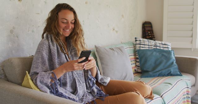 Young woman sitting on a comfortable couch, smiling while using her smartphone. She looks relaxed and cozy in casual attire. Can be used for themes related to technology, home lifestyle, communication, social media engagement, or depicting a comfortable and modern living environment.