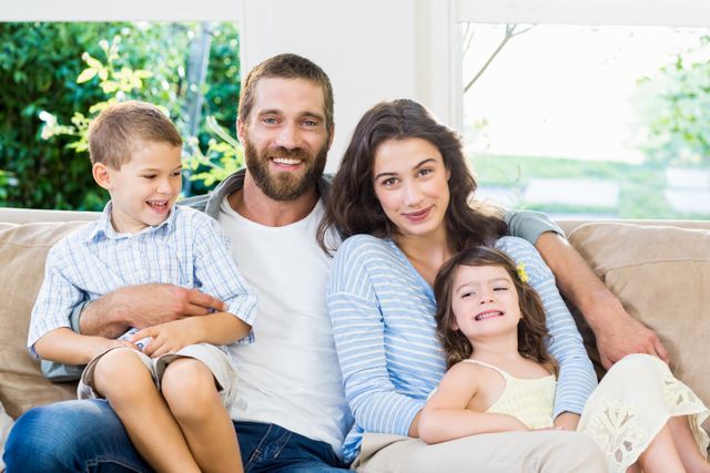 A happy family, including parents and two young children, sitting closely on a sofa in a bright and modern living room. Their expressions are joyful, reflecting love and togetherness. This image can be used for advertisements, family-oriented campaigns, articles on parenting, and lifestyle blogs. It showcases a relaxed and loving family environment, ideal for promoting family-related products and services.