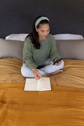 Mid adult asian woman sitting on bed journaling and using smartphone in cozy bedroom