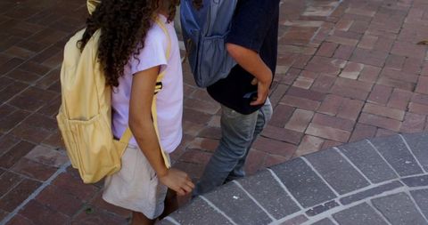 Two schoolchildren walking on sunlit brick courtyard carrying yellow and light blue backpacks
