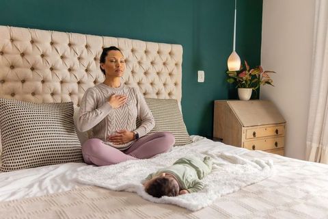 Mid-30s mother practicing mindful breathing on bed with sleeping infant daughter