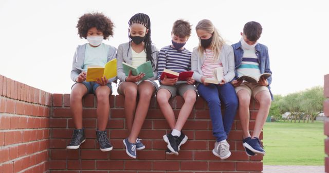 Group of children sitting on brick wall, reading books. Children wearing masks for safety while reading books outdoors. Can be used for education, safety, diversity, and children's activities themes.