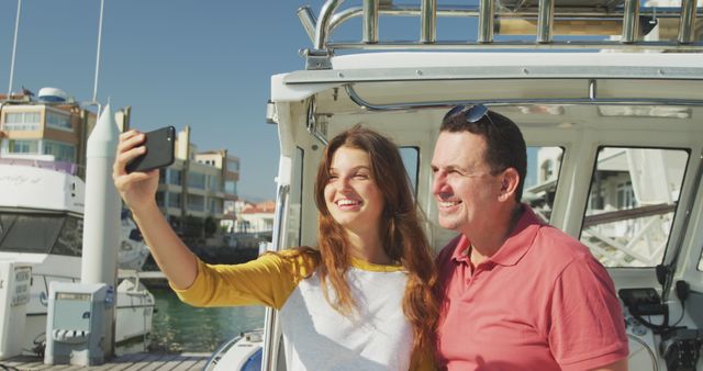 Father and daughter taking a selfie together while standing on a boat at a marina. Both are smiling and enjoying their time under the sunshine, showcasing a special bonding moment. Suitable for promoting family vacations, outdoor activities, and travel experiences. Ideal for articles and adverts focusing on family bonding, summer holidays, and parent-child relationships.