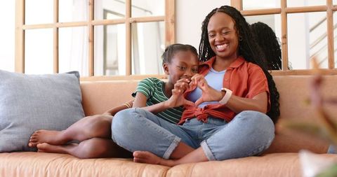 African american mother and daughter sharing heart gesture on living room couch, cozy bond