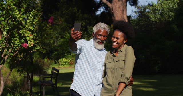 Heartwarming moment of family bonding. Senior father and young daughter are taking a selfie in a lush garden. Ideal for use in articles about family relationships, leisure activities, technology integration in daily life, and outdoor enjoyment. Great for promoting family events, mobile phone cameras, or lifestyle blogs with a focus on family and nature.