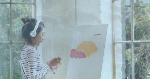Artist painting at easel with headphones near rain-streaked window in bright natural light