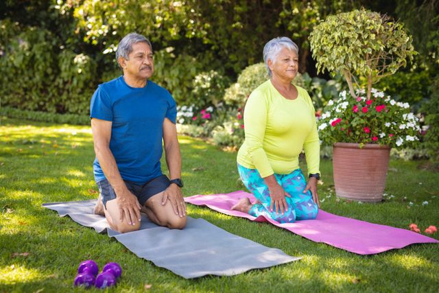 Biracial senior couple looking away while kneeling on exercise mats against plants in park. nature, yoga, exercise, unaltered, love, togetherness, retirement, fitness and active lifestyle concept.