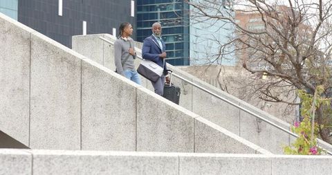 Senior Businessman Walking Down Urban Staircase with Young Colleague Pulling Suitcase