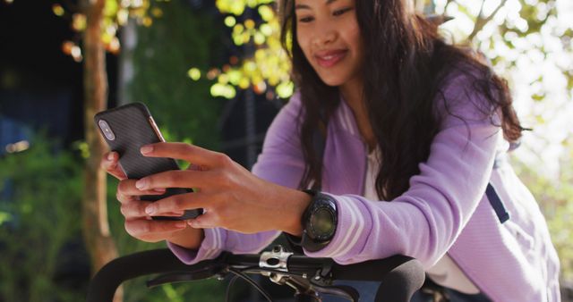 A young Asian woman is taking a break while cycling in the countryside, using her smartphone. She is dressed in a purple casual jacket and seems relaxed while interacting with her phone. The background is filled with autumnal trees and greenery, contributing to a serene atmosphere. This could be used for themes of technology in daily life, connected lifestyle, outdoor leisure activities, or autumn experiences.