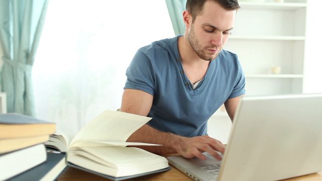 Young man working on a laptop at a desk, surrounded by open books, in home environment with soft natural lighting. Useful for depicting studying or working from home, online education, and independent learning concepts.