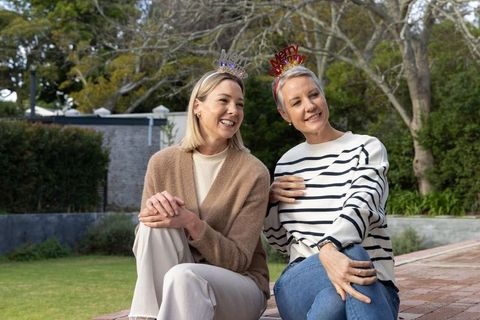 Two women sitting in backyard wearing merry christmas headbands, laughing together, cozy