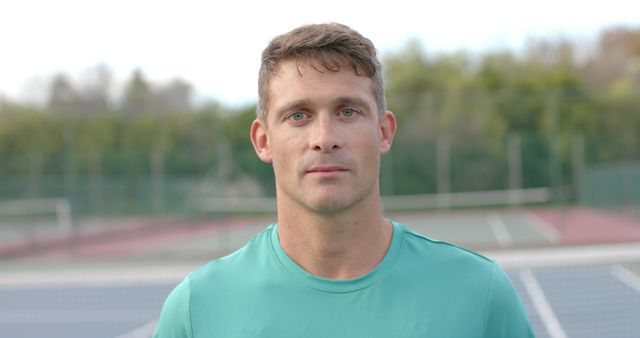 Middle-aged male tennis player standing on an outdoor court, wearing a green shirt and looking directly at the camera with confident expression. Ideal for articles on sports, fitness coaching, tennis training, or promotion of sports apparel and equipment.