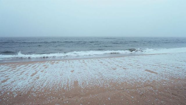 Misty shoreline with sea foam lapping wet ripple sand and gentle waves on overcast coast