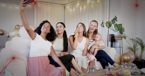 Women taking selfie on couch at baby shower celebration with balloons and fairy lights