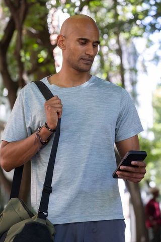 African american man checking smartphone on sunlit urban path with shoulder bag