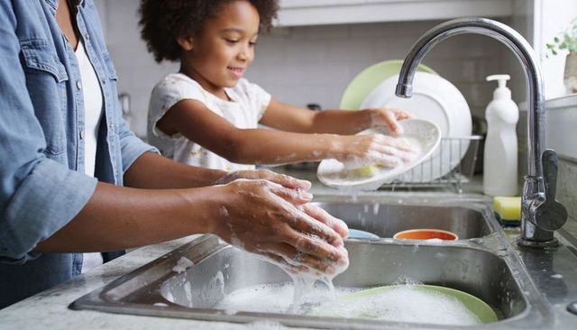 Parent and child washing dishes together at kitchen sink teaching responsibility, bonding