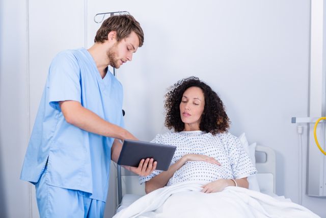 Medical professional assisting pregnant woman using digital tablet in hospital ward. Ideal for healthcare, maternity, and technology in healthcare themes. Useful for illustrating patient care, medical consultations, and hospital environments.