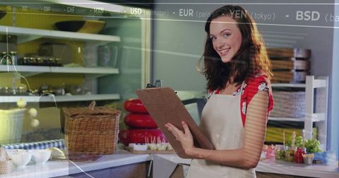 Smiling shop assistant checking clipboard beside stacked red cheeses at deli counter wearing apron