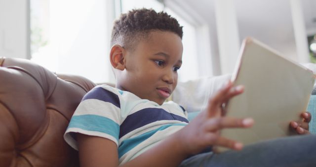 Young boy engrossed with a tablet while lounging on a brown leather sofa at home. Ideal for use in campaigns focusing on children's technology use, educational content, e-learning setups, or portraying home relaxation and contemporary family life.
