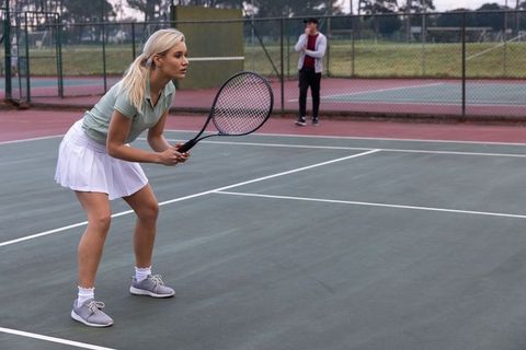 Female tennis player crouching in ready stance on outdoor hard court with spectator observing
