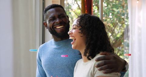 Laughing couple sharing warm moment by sunlit glass door with UI label overlays, cozy vibe