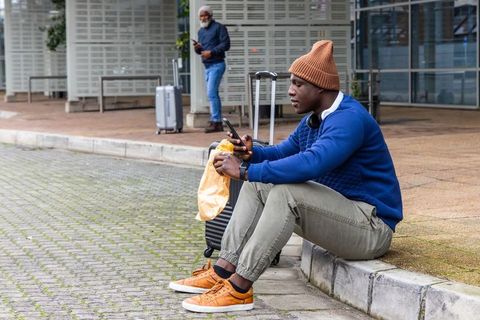 Traveler sitting on curb checking phone with suitcase and takeout while waiting in transit
