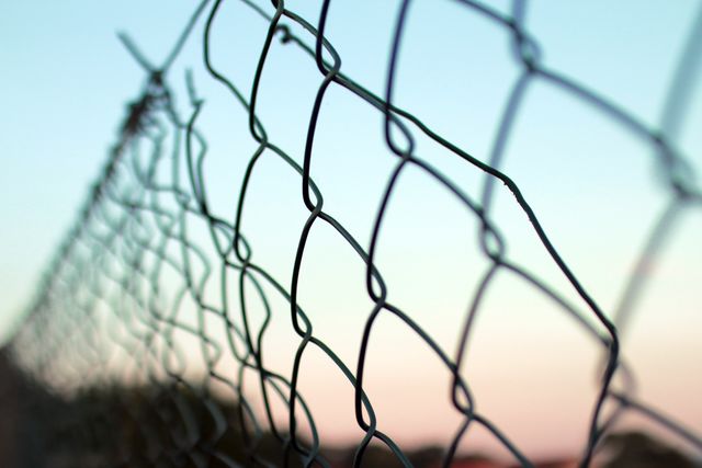 Capturing a detailed view of a chain-link fence against a backdrop of a serene sunset. This composition highlights the intricate weaving of the fence and the gradient sky, evoking themes of security contrasted with freedom. Useful for illustrating concepts of barriers, boundaries, protection, and urbanization in outdoor settings. Ideal for blogs, presentations, and articles discussing these abstract concepts or for use in security-related content.