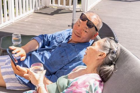 Relaxing mature couple lounging on sunlit patio deck, sharing smartphone and drinks