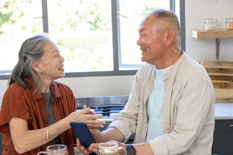 Senior asian couple sharing warm conversation in modern kitchen while holding smartphone