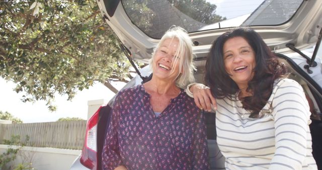 Two mature women are laughing and enjoying each other's company near a car in an outdoor setting. Ideal for concepts relating to friendship, retirement, leisure time, road trips, and outdoor activities. Can be used in advertisements for travel, senior lifestyle, and happy moments.