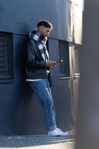 Young man leaning on urban wall checking phone wearing checked scarf and leather jacket