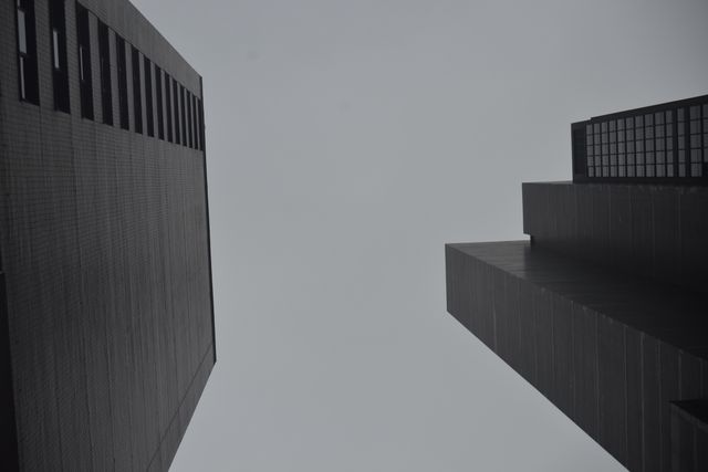 View of modern city skyscrapers from below looking upwards, showcasing minimalist perspective. Dark, overcast sky enhances cold, moody atmosphere. Useful for illustrating city life, business concepts, urban development, contemporary architecture studies or minimalistic design themes.