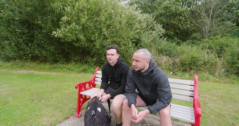 Two men resting on park bench after jogging in lush green park