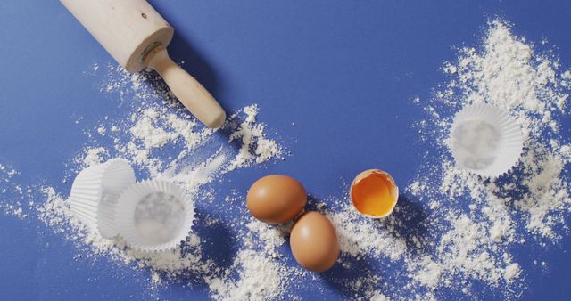 Image of baking ingredients, muffin papers, eggs and tools lying on blue surface with flour. baking, food preparing, taste and flavour concept.