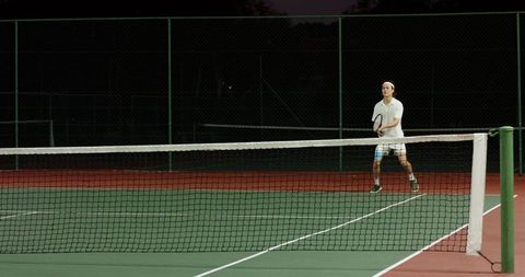 Asian man wearing polo and headband practicing tennis at night on outdoor court
