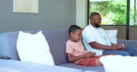 African american father and son playing video games together on living room sofa