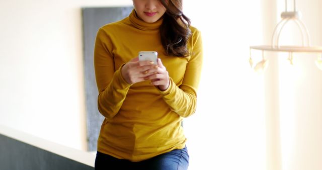 Young woman standing indoors, engaging with smartphone. Ideal for technology, communication, social media, smartphone usage, and modern lifestyle themes.
