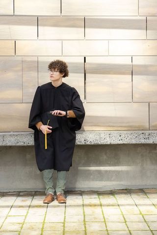 Young male graduate leaning on stone ledge holding mortarboard and smartphone