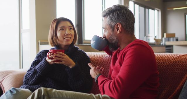 Man and woman sitting on couch, enjoying hot drinks and conversation. Perfect for ads or articles on lifestyle, relaxation, home comfort, and relationships. Ideal for use in promotional material for cozy living and beverages.