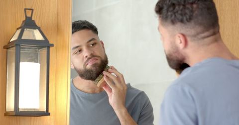 Man grooming beard with wooden brush in bathroom mirror reflection