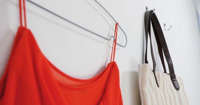 A red fabric bag and a beige tote with black straps hang on silver hooks against a white wall, with copy space. Such an arrangement is often found in a home or office setting, providing a neat storage solution for personal items.