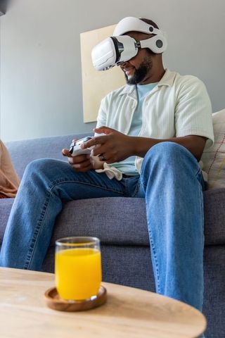 African american man playing vr game on couch holding controller and glass of orange juice