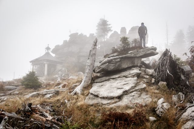 Mysterious and adventurous scene of a person standing on rocky terrain surrounded by fog in a mountain landscape with scattered twisted, dry trees and a gazebo structure in the background. Ideal for themes focusing on exploration, travel, and the tranquility of nature.