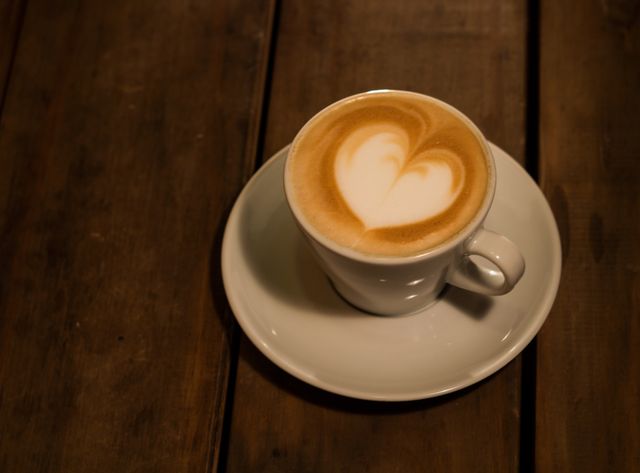 Heart-shaped latte art in a white cup placed on a rustic wooden table, ideal for illustrating cozy cafes, coffee shops, breakfast themes, or barista skills.